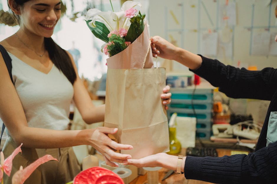 A woman with long dark hair, wearing a white sleeveless top, is smiling while receiving a bouquet of fresh flowers from a person dressed in black. The bouquet, wrapped in pink paper and placed inside