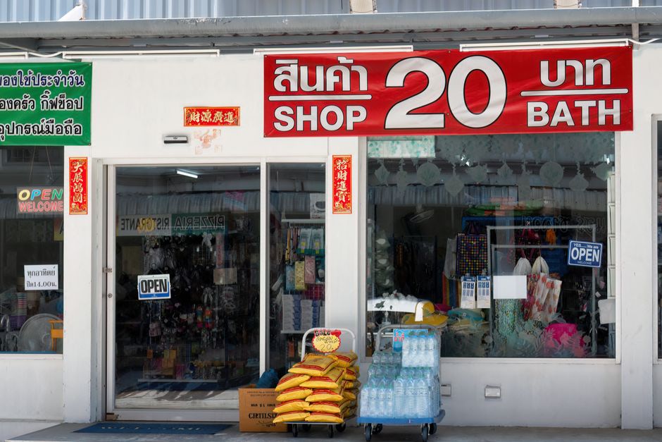 A storefront of a shop offering flowers and other goods, with a large red sign displaying Thai characters and the words 'SHOP 20 BATH' in white. The shop has glass doors and windows revealing various 