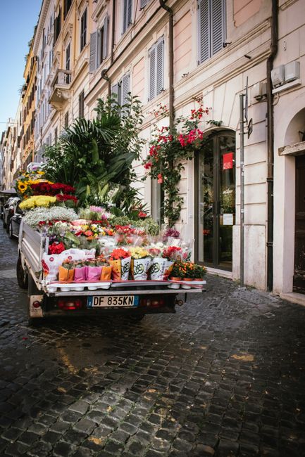A flower delivery truck parked on a cobblestone street in front of a building with light-colored stone walls and large windows, decorated with blooming pink and red flowers hanging from window boxes.