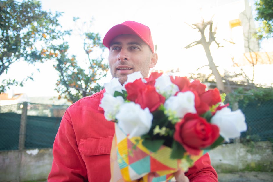 A male flower delivery courier from flowerdeliverywinchmorehill.co.uk is outdoors in natural daylight, wearing a red cap and matching red jacket, holding a bouquet of fresh flowers. The bouquet featur