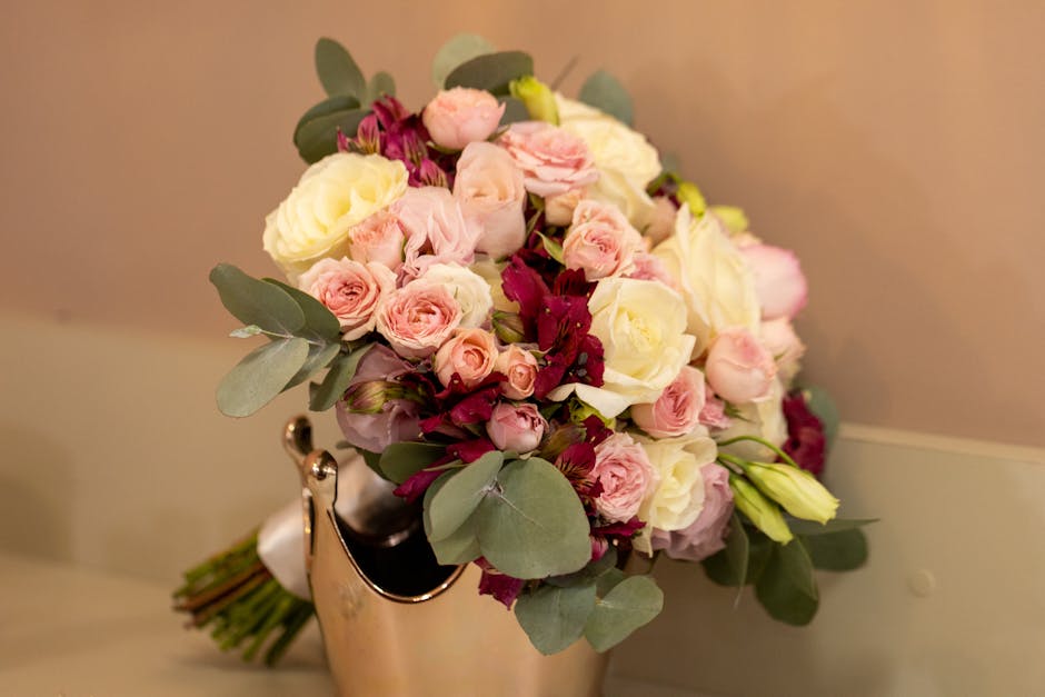 A bouquet of fresh flowers arranged in a rounded shape, featuring soft pink roses, creamy white roses, and cream-colored lisianthus, accented with dark red alstroemeria and silvery-green eucalyptus le
