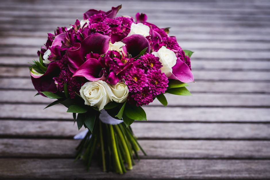 A vibrant bouquet featuring deep magenta calla lilies, bright pink chrysanthemums, and rich purple blooms arranged with white roses, complemented by fresh green foliage. The flowers are tightly cluste